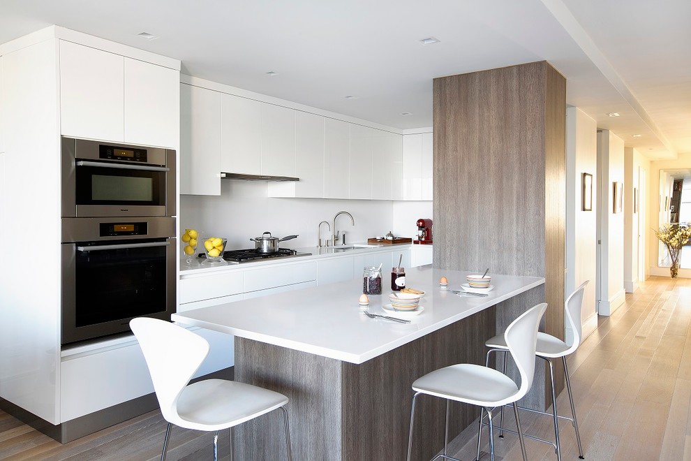 Example of a trendy l-shaped light wood floor kitchen design in New York with an undermount sink, flat-panel cabinets, white cabinets, white backsplash, stainless steel appliances and a peninsula