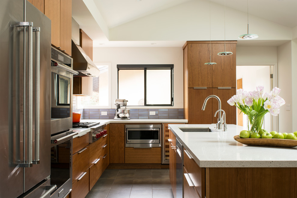 Modern inredning av ett l-kök, med en undermonterad diskho, släta luckor, skåp i mellenmörkt trä, grått stänkskydd, rostfria vitvaror och en köksö
