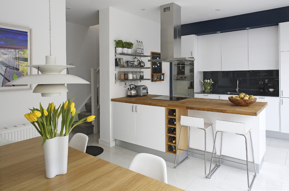 Modelo de cocina comedor actual con electrodomésticos de acero inoxidable, encimera de madera, armarios con paneles lisos, puertas de armario blancas, salpicadero negro, salpicadero de vidrio templado y encimeras marrones