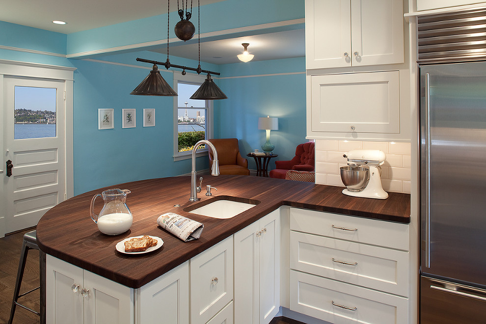 Foto på ett mellanstort funkis kök, med rostfria vitvaror, en rustik diskho, skåp i shakerstil, vita skåp, träbänkskiva, vitt stänkskydd, stänkskydd i tunnelbanekakel, mörkt trägolv, en halv köksö och brunt golv