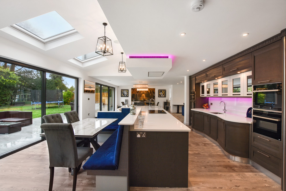 Expansive contemporary galley kitchen/diner in London with a submerged sink, shaker cabinets, medium wood cabinets, white splashback, stainless steel appliances, an island, brown floors and beige worktops.