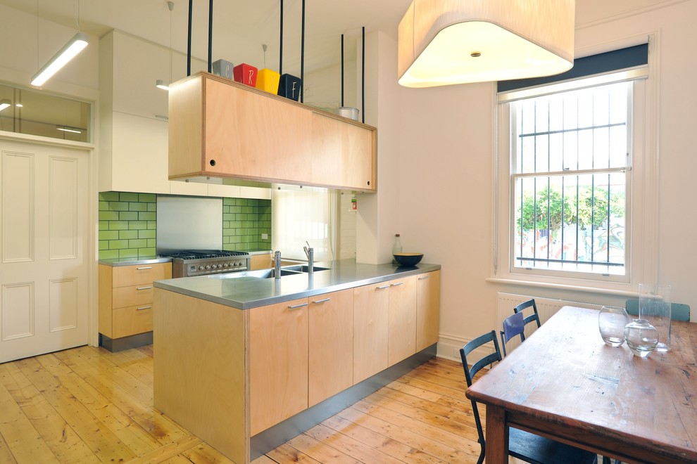 Foto på ett funkis kök, med skåp i ljust trä, grönt stänkskydd och rostfria vitvaror