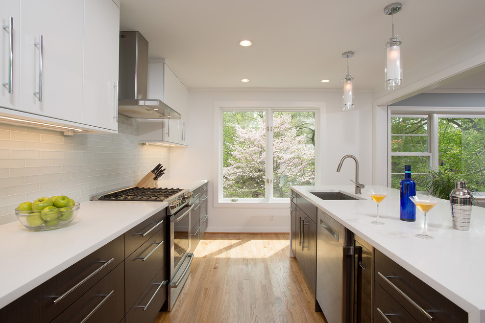 This is an example of a medium sized contemporary u-shaped open plan kitchen in DC Metro with a submerged sink, flat-panel cabinets, white cabinets, composite countertops, white splashback, metro tiled splashback, stainless steel appliances, light hardwood flooring and an island.