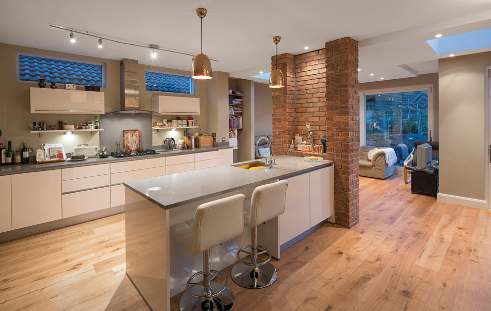 Inspiration for a contemporary single-wall open plan kitchen in Dublin with a submerged sink, flat-panel cabinets, white cabinets, composite countertops, grey splashback, stainless steel appliances, light hardwood flooring and a breakfast bar.