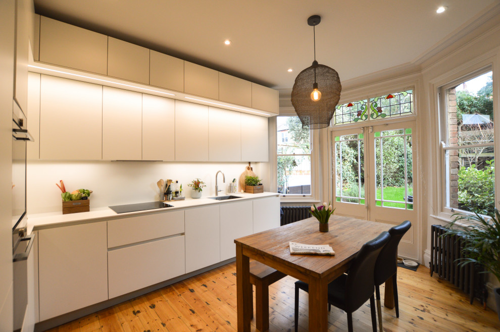 Photo of a medium sized contemporary l-shaped kitchen/diner in London with flat-panel cabinets, white cabinets, quartz worktops, stainless steel appliances, no island, white worktops, a submerged sink, medium hardwood flooring and brown floors.