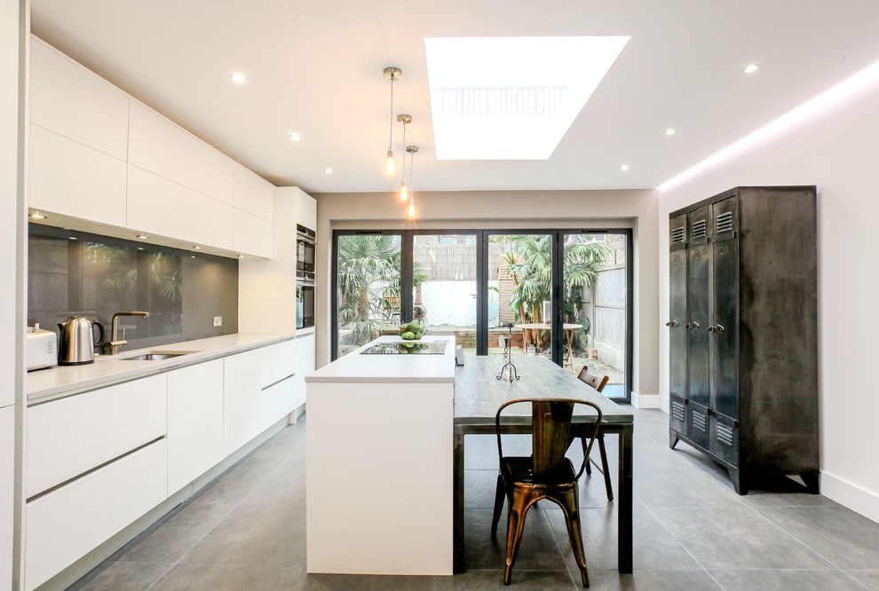 Large contemporary galley kitchen/diner in London with a submerged sink, flat-panel cabinets, white cabinets, grey splashback, glass sheet splashback, black appliances, an island, composite countertops and ceramic flooring.