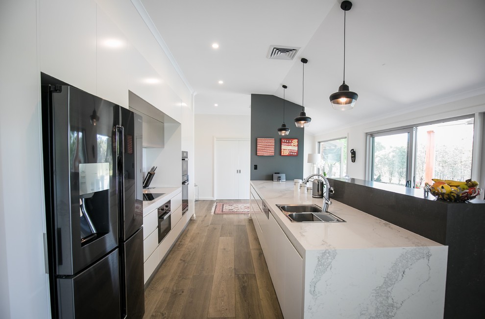 Photo of a contemporary galley kitchen in Sydney with a built-in sink, flat-panel cabinets, white cabinets, black appliances, medium hardwood flooring, a breakfast bar, brown floors and grey worktops.