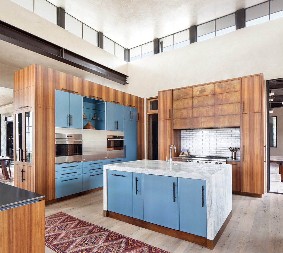 Foto på ett funkis kök, med släta luckor, blå skåp, vitt stänkskydd, stänkskydd i tunnelbanekakel, rostfria vitvaror, ljust trägolv och en köksö