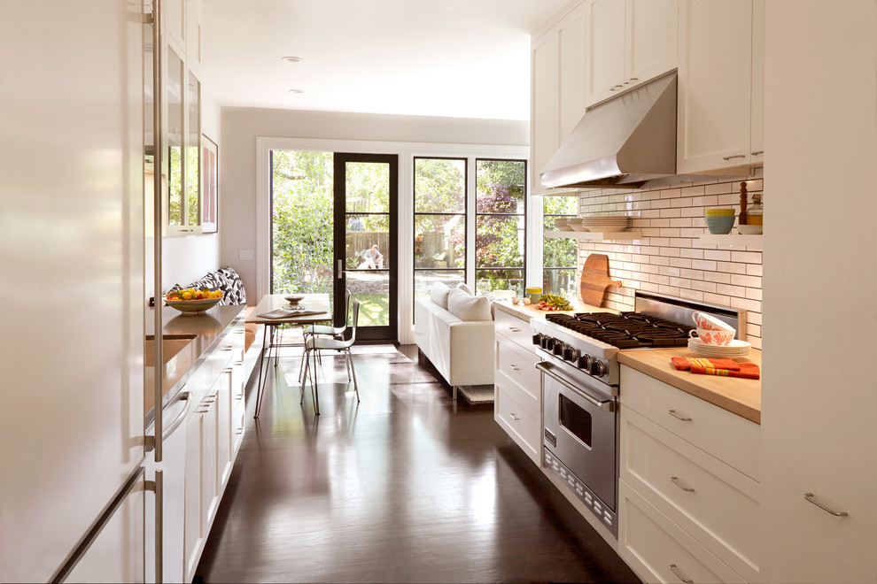 Diseño de cocina comedor actual de tamaño medio sin isla con puertas de armario blancas, salpicadero blanco, electrodomésticos de acero inoxidable, suelo de madera oscura, armarios con paneles lisos, encimera de madera, salpicadero de azulejos tipo metro, fregadero bajoencimera y suelo marrón