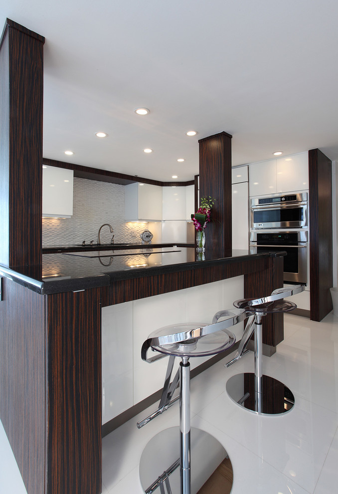 Example of a large trendy l-shaped porcelain tile and white floor eat-in kitchen design in Miami with stainless steel appliances, an island, a drop-in sink, flat-panel cabinets, white cabinets, solid surface countertops, white backsplash and black countertops