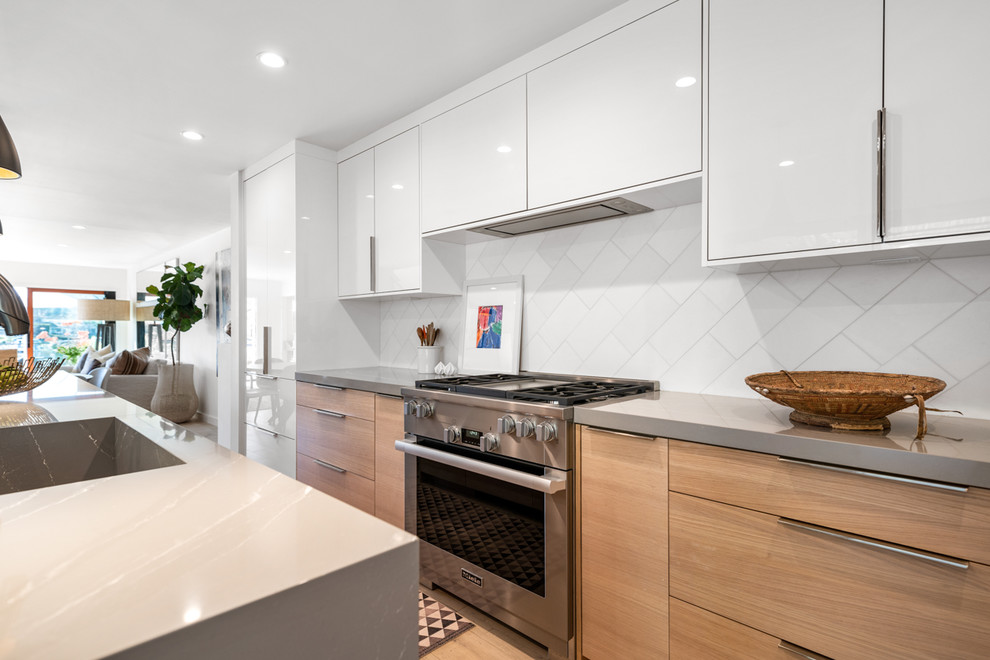 This is an example of a large contemporary open plan kitchen in Los Angeles with an integrated sink, flat-panel cabinets, white cabinets, engineered stone countertops, white splashback, marble splashback, stainless steel appliances, light hardwood flooring, an island, beige floors and grey worktops.
