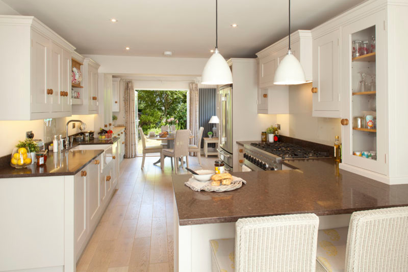 This is an example of a large traditional l-shaped open plan kitchen in Kent with a belfast sink, shaker cabinets, grey cabinets, granite worktops, glass sheet splashback, stainless steel appliances, light hardwood flooring and a breakfast bar.