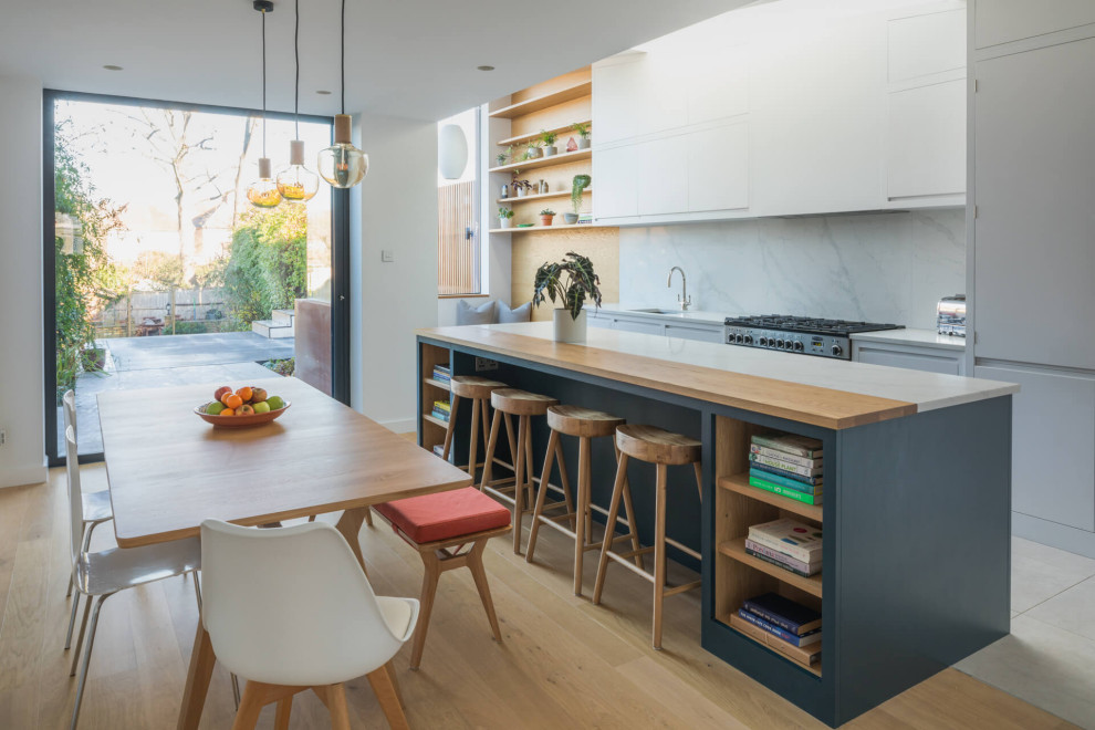 Large contemporary single-wall open plan kitchen in London with a built-in sink, flat-panel cabinets, white cabinets, quartz worktops, white splashback, stone slab splashback, stainless steel appliances, ceramic flooring, an island, white floors and white worktops.