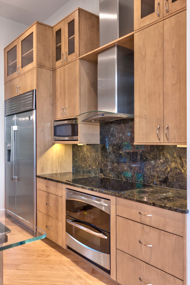 Cette image montre une cuisine design en bois clair avec un placard à porte plane, un plan de travail en verre, une crédence en dalle de pierre et un électroménager en acier inoxydable.