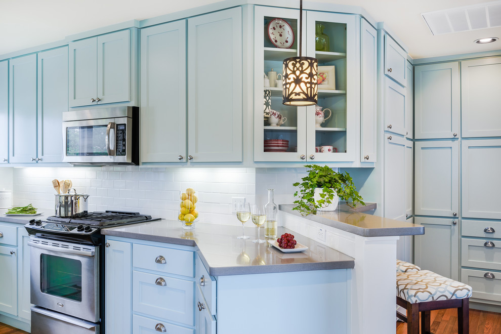 Cette image montre une cuisine américaine vintage en U avec un évier de ferme, un placard à porte shaker, des portes de placard bleues, un plan de travail en surface solide, une crédence blanche, une crédence en carrelage métro, un électroménager en acier inoxydable, un sol en bois brun et aucun îlot.