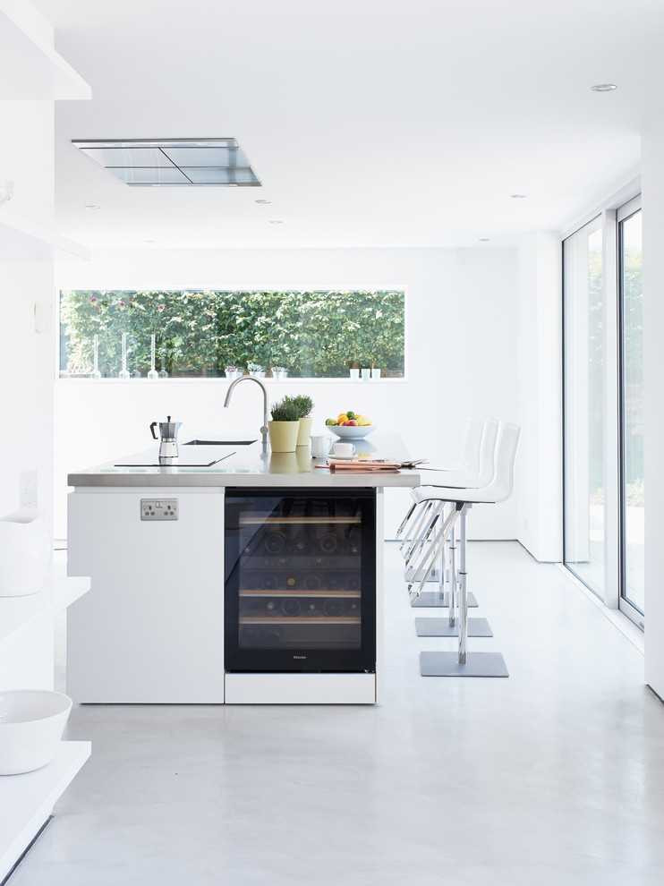 Große, Einzeilige Moderne Wohnküche mit Kücheninsel, flächenbündigen Schrankfronten, weißen Schränken, Edelstahl-Arbeitsplatte, schwarzen Elektrogeräten, integriertem Waschbecken und Betonboden in Sussex