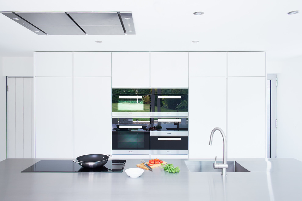 Diseño de cocina comedor lineal contemporánea grande con una isla, armarios con paneles lisos, puertas de armario blancas, encimera de acero inoxidable, electrodomésticos negros, fregadero integrado y suelo de cemento
