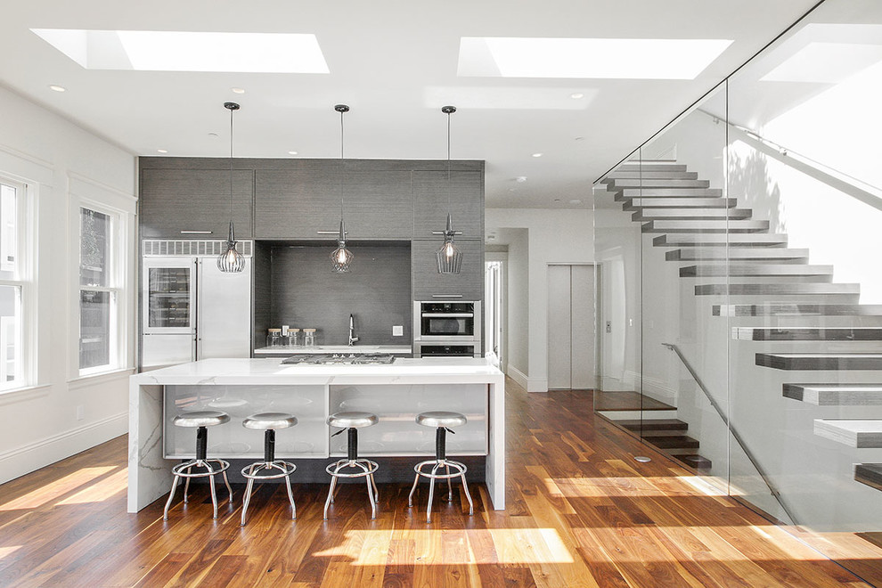 Medium sized contemporary single-wall open plan kitchen in San Francisco with flat-panel cabinets, dark wood cabinets, black splashback, stainless steel appliances, an island, a submerged sink, marble worktops, dark hardwood flooring, brown floors and white worktops.