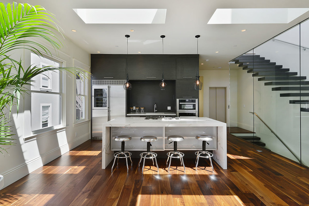 Inspiration for a medium sized contemporary single-wall open plan kitchen in San Francisco with flat-panel cabinets, dark wood cabinets, black splashback, stainless steel appliances, an island, a submerged sink, marble worktops, dark hardwood flooring, brown floors and white worktops.