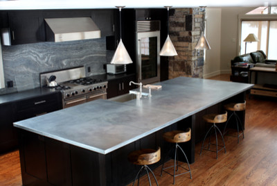 Mid-sized trendy single-wall medium tone wood floor open concept kitchen photo in Denver with gray backsplash, stone slab backsplash, stainless steel appliances, an island, a farmhouse sink, shaker cabinets, black cabinets and concrete countertops