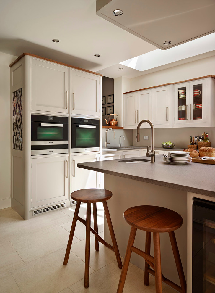 Medium sized contemporary l-shaped open plan kitchen in London with shaker cabinets, stainless steel appliances, an island, a built-in sink, grey cabinets, grey splashback, glass sheet splashback and limestone flooring.