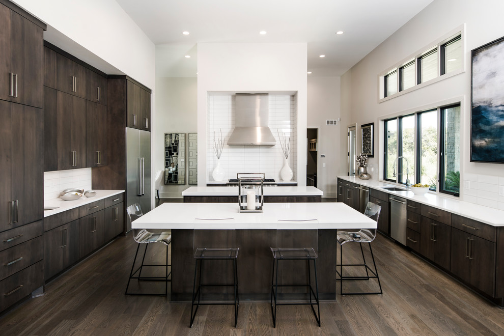 Ejemplo de cocina actual con fregadero bajoencimera, armarios con paneles lisos, puertas de armario de madera en tonos medios, salpicadero blanco, electrodomésticos de acero inoxidable, suelo de madera oscura, dos o más islas, suelo marrón y encimeras blancas