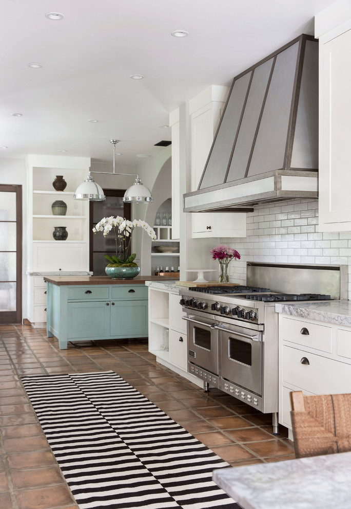 ロサンゼルスにある地中海スタイルのおしゃれなアイランドキッチン (フラットパネル扉のキャビネット、白いキャビネット、白いキッチンパネル、サブウェイタイルのキッチンパネル、シルバーの調理設備、テラコッタタイルの床) の写真