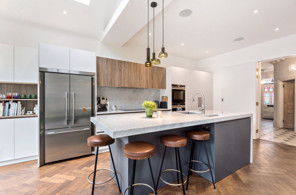 Example of a trendy medium tone wood floor kitchen design in London with flat-panel cabinets, marble countertops, gray backsplash, marble backsplash, stainless steel appliances, an island, gray countertops, an undermount sink and white cabinets