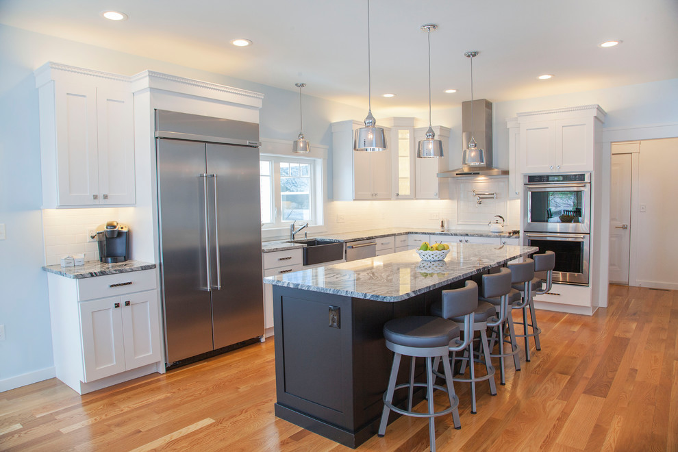 Example of a large trendy l-shaped light wood floor and brown floor eat-in kitchen design in Boston with a farmhouse sink, shaker cabinets, white cabinets, granite countertops, white backsplash, subway tile backsplash, stainless steel appliances and an island