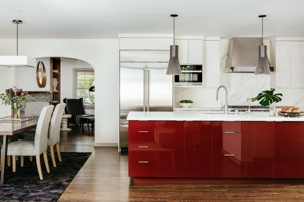 Inredning av ett modernt vit vitt kök, med en undermonterad diskho, släta luckor, röda skåp, vitt stänkskydd, rostfria vitvaror, mellanmörkt trägolv, en köksö och brunt golv