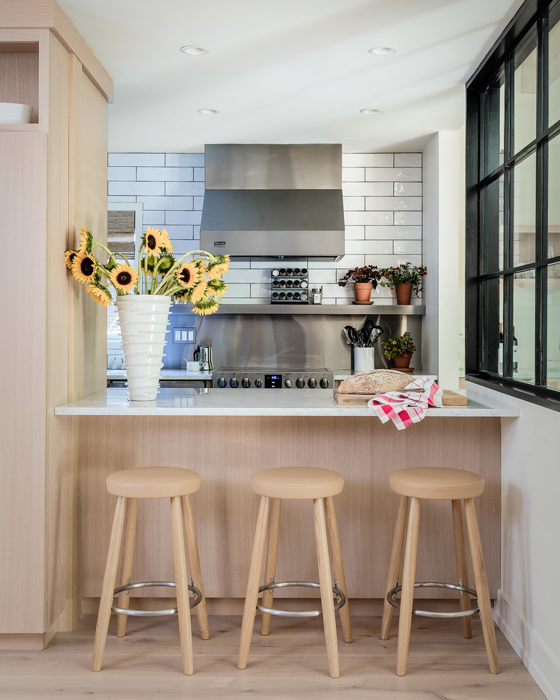 На фото: параллельная кухня в современном стиле с светлыми деревянными фасадами, техникой из нержавеющей стали, белой столешницей, белым фартуком, светлым паркетным полом, полуостровом, бежевым полом и фартуком из плитки кабанчик