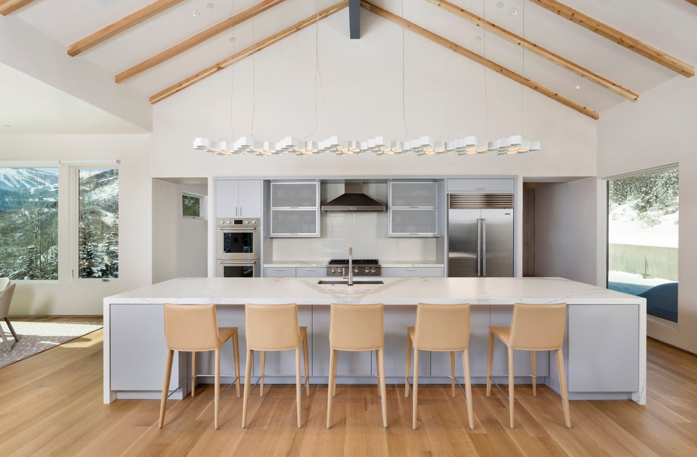 Foto på ett mellanstort funkis linjärt kök med öppen planlösning, med en undermonterad diskho, vitt stänkskydd, stänkskydd i glaskakel, rostfria vitvaror, ljust trägolv, en köksö, släta luckor, grå skåp, marmorbänkskiva och brunt golv