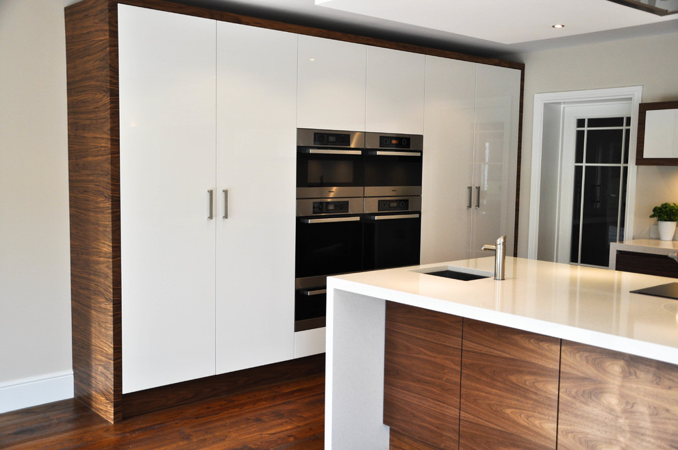 This is an example of a large contemporary l-shaped open plan kitchen in Surrey with a built-in sink, flat-panel cabinets, dark wood cabinets, quartz worktops, stainless steel appliances, dark hardwood flooring and an island.