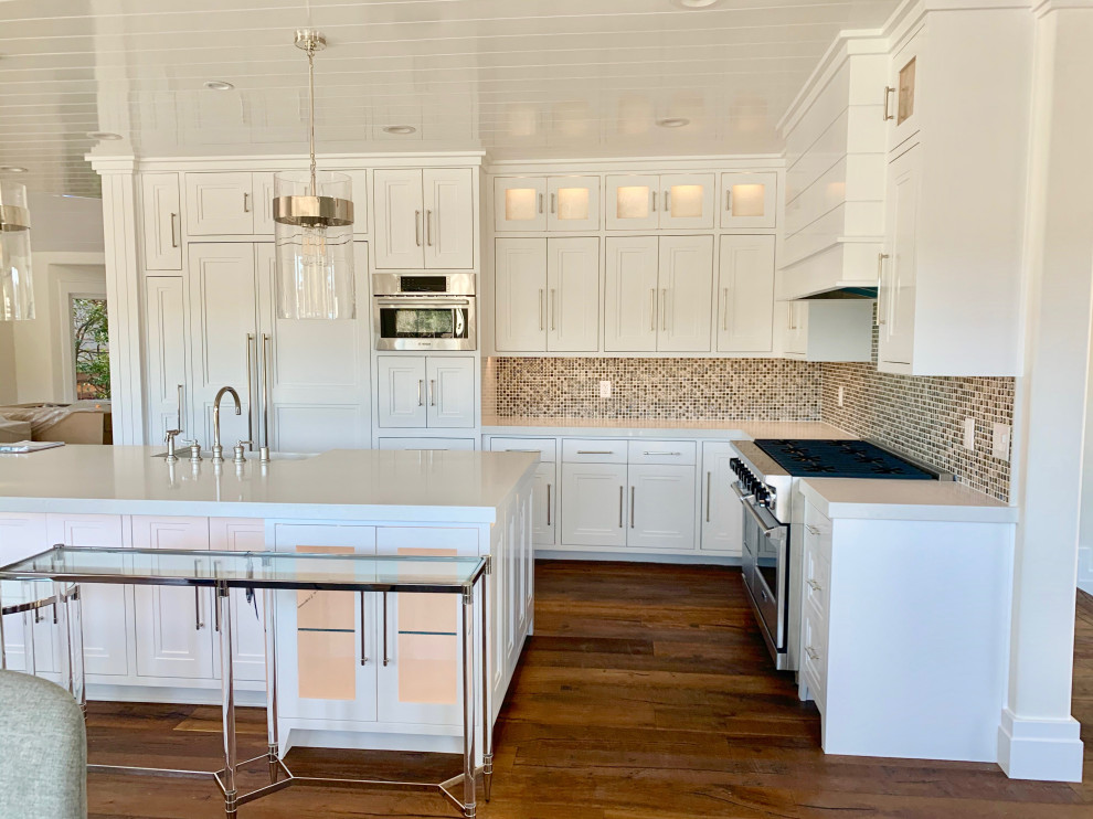 Modern inredning av ett stort vit vitt kök, med en rustik diskho, luckor med profilerade fronter, vita skåp, bänkskiva i kvarts, brunt stänkskydd, stänkskydd i mosaik, rostfria vitvaror, mörkt trägolv, en köksö och brunt golv