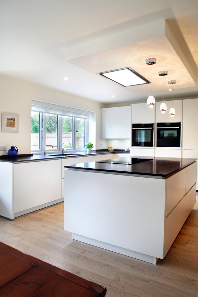 Example of a mid-sized trendy l-shaped medium tone wood floor eat-in kitchen design in Buckinghamshire with flat-panel cabinets, white cabinets, quartzite countertops, an island, an integrated sink and black appliances