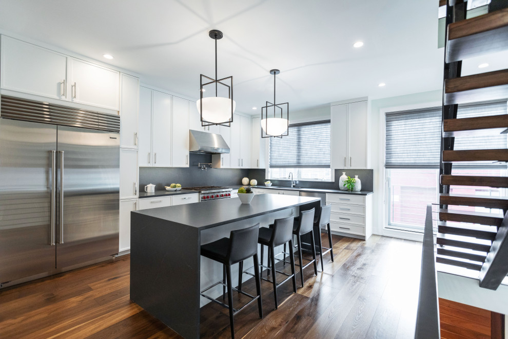 This is an example of a contemporary l-shaped enclosed kitchen in Philadelphia with recessed-panel cabinets, white cabinets, grey splashback, stone slab splashback, stainless steel appliances, dark hardwood flooring, an island, brown floors and grey worktops.