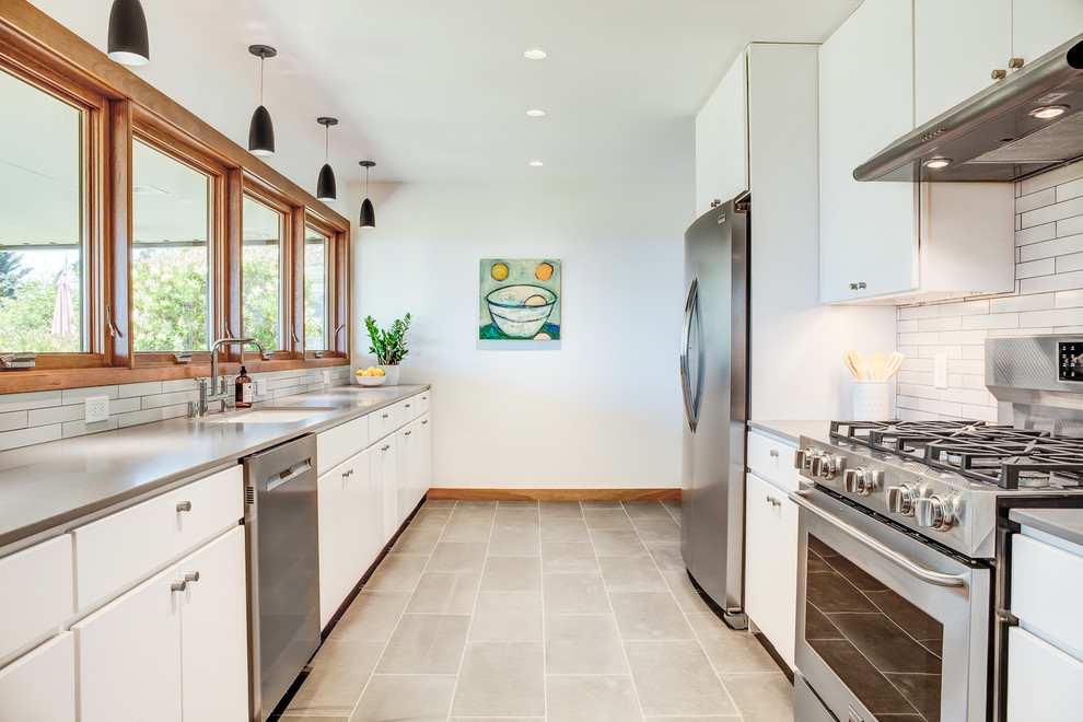 サンフランシスコにある広いコンテンポラリースタイルのおしゃれなキッチン (アンダーカウンターシンク、フラットパネル扉のキャビネット、白いキャビネット、ステンレスカウンター、白いキッチンパネル、シルバーの調理設備、アイランドなし、グレーの床、グレーのキッチンカウンター、サブウェイタイルのキッチンパネル、コンクリートの床) の写真