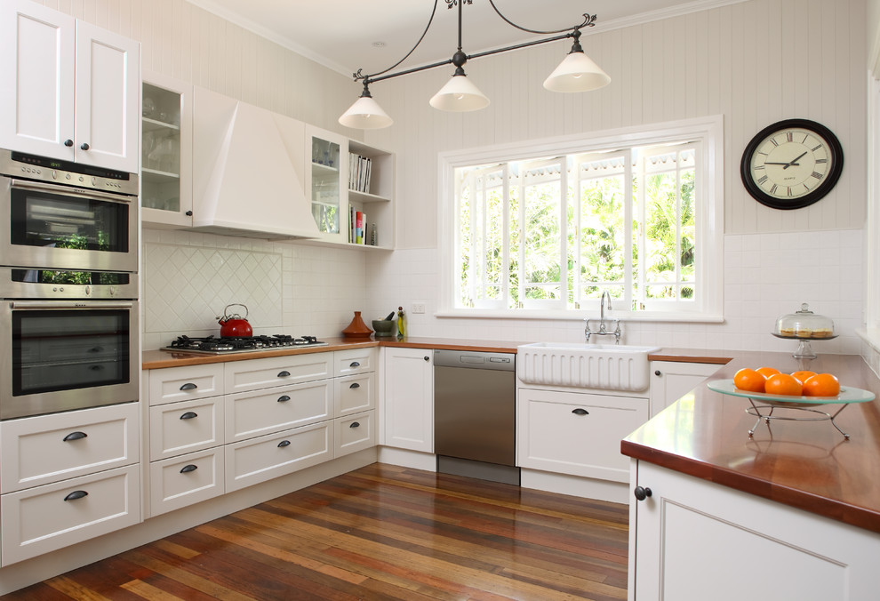 Klassisk inredning av ett kök, med rostfria vitvaror, en rustik diskho, luckor med infälld panel, vita skåp, vitt stänkskydd och träbänkskiva