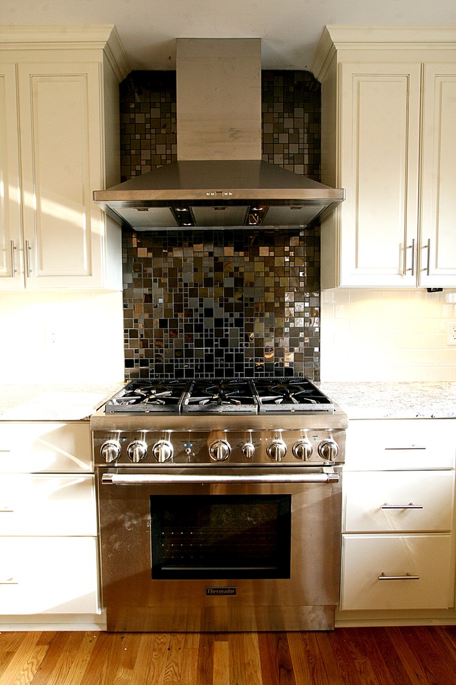 Cette image montre une cuisine design avec des portes de placard blanches, un plan de travail en granite, une crédence noire, un sol en bois brun et îlot.