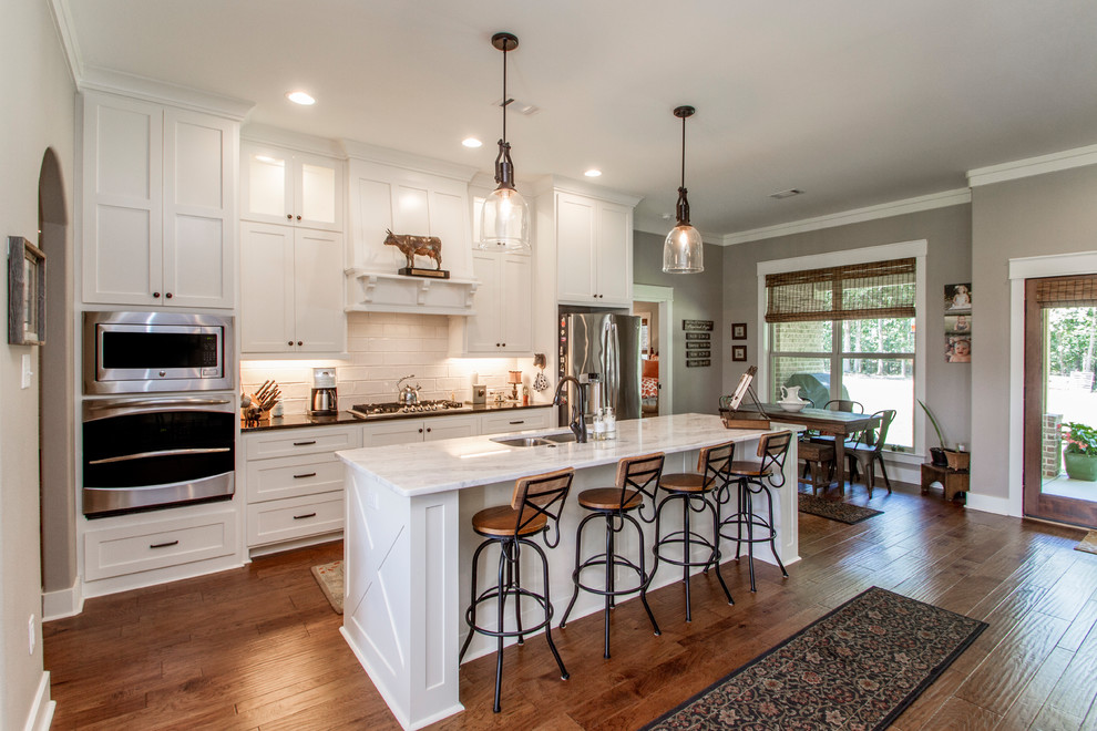 Mid-sized country galley medium tone wood floor and brown floor open concept kitchen photo in Little Rock with a double-bowl sink, shaker cabinets, white cabinets, quartz countertops, white backsplash, ceramic backsplash, stainless steel appliances, an island and white countertops