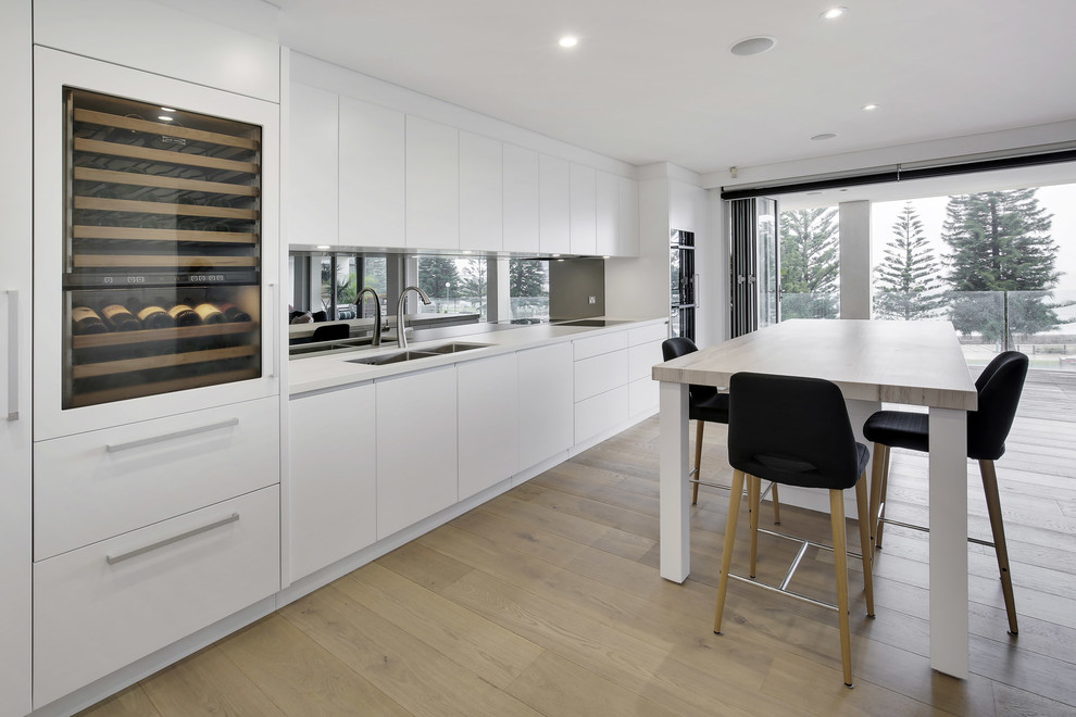 Inspiration for a large scandi l-shaped open plan kitchen in Sydney with a submerged sink, flat-panel cabinets, white cabinets, engineered stone countertops, metallic splashback, mirror splashback, black appliances, light hardwood flooring, an island and yellow floors.