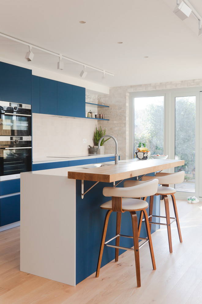 Photo of a medium sized contemporary galley kitchen/diner in London with a double-bowl sink, flat-panel cabinets, blue cabinets, beige splashback, ceramic splashback, integrated appliances, medium hardwood flooring, an island and beige floors.