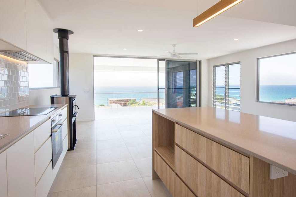 This is an example of a medium sized beach style l-shaped open plan kitchen in Brisbane with flat-panel cabinets, white cabinets, white splashback, an island, beige floors, metro tiled splashback, stainless steel appliances and ceramic flooring.