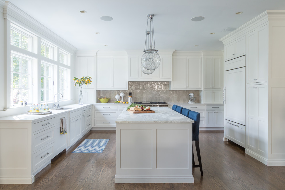 Inredning av ett klassiskt u-kök, med en undermonterad diskho, luckor med infälld panel, vita skåp, grått stänkskydd, integrerade vitvaror, mörkt trägolv, en köksö och brunt golv