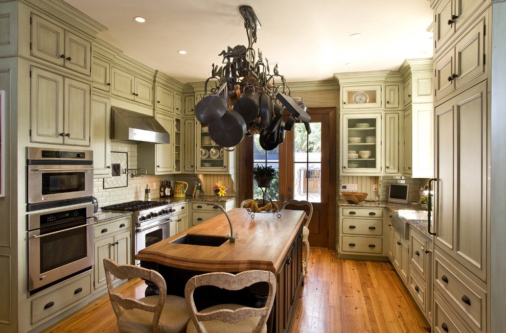 Example of a large arts and crafts u-shaped medium tone wood floor and brown floor enclosed kitchen design in Charleston with a farmhouse sink, light wood cabinets, wood countertops, subway tile backsplash, paneled appliances, beaded inset cabinets, white backsplash, an island and brown countertops