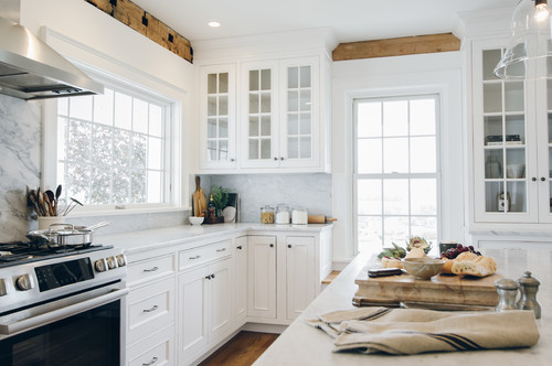 Bright white kitchen with glass-front RTA shaker cabinets, marble backsplash and countertops, stainless range, and large windows accented by rustic wood beams.