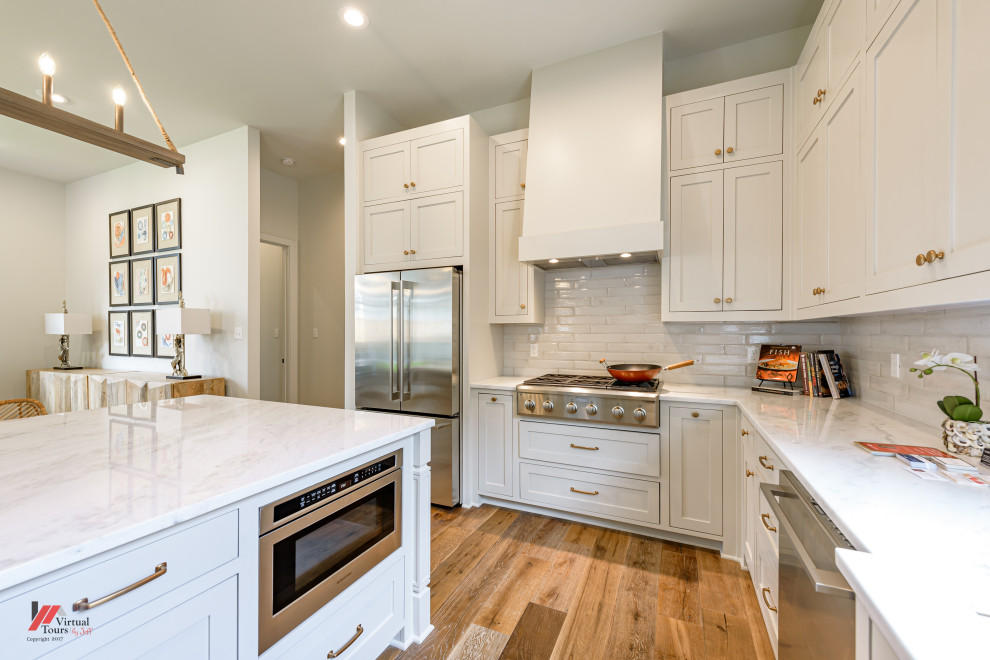 Inspiration for a large traditional l-shaped kitchen in New Orleans with white cabinets, white splashback, metro tiled splashback, stainless steel appliances, medium hardwood flooring, an island, white worktops, shaker cabinets and brown floors.