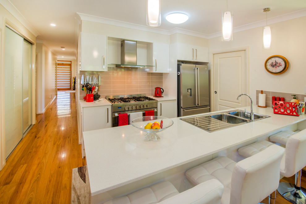 Example of a transitional galley medium tone wood floor kitchen design in Sydney with a drop-in sink, flat-panel cabinets, white cabinets, beige backsplash, ceramic backsplash, stainless steel appliances and a peninsula