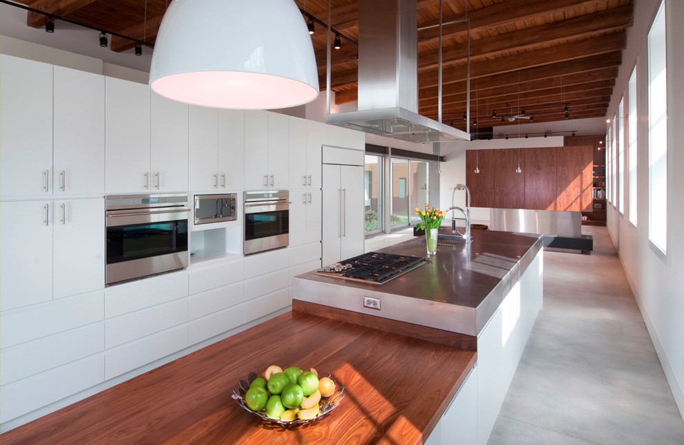 Immagine di un cucina con isola centrale minimalista con lavello sottopiano, ante lisce, ante bianche, top in legno, elettrodomestici in acciaio inossidabile e pavimento in cemento