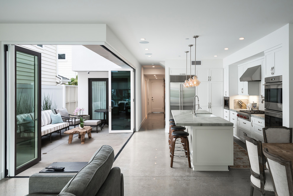 Mid-sized transitional l-shaped concrete floor and gray floor open concept kitchen photo in Orange County with a farmhouse sink, shaker cabinets, white cabinets, white backsplash, stainless steel appliances, an island, marble countertops, subway tile backsplash and gray countertops